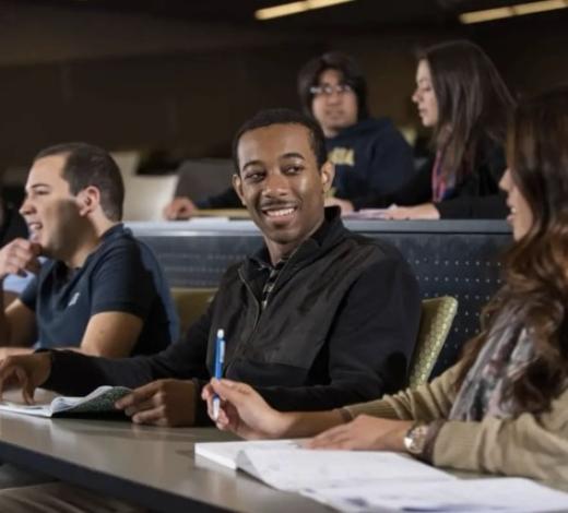Photo of an African American male student talk to a white female student sitting at an auditorium desk while other students behind them look to the front of the room 
