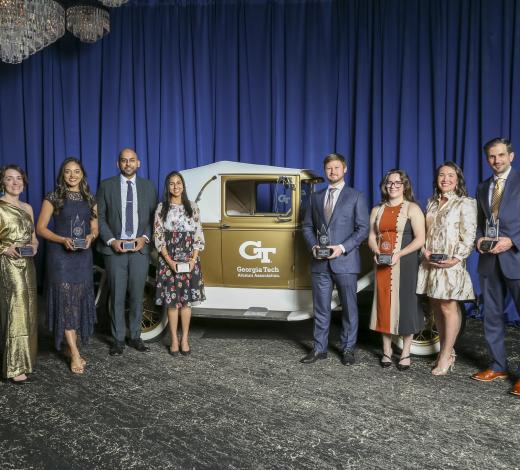 A group of people stand on a stage in front of a vintage Georgia Tech Ramblin’ Wreck car, each holding a glass award, with blue draping and chandeliers in the background.