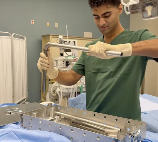 A person wearing surgical scrubs and gloves uses a metal tool to work with a stainless‑steel medical device or instrument tray in a clinical environment. Medical equipment and curtains are visible in the background.