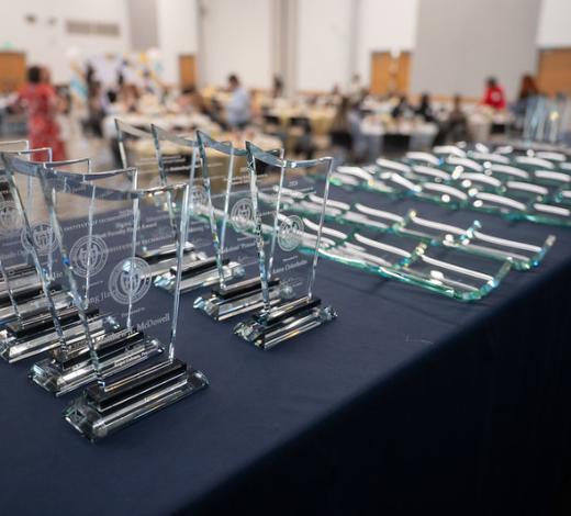 A row of clear glass award plaques arranged on a table in the foreground, with more plaques laid flat beside them and a blurred event setup in the background.