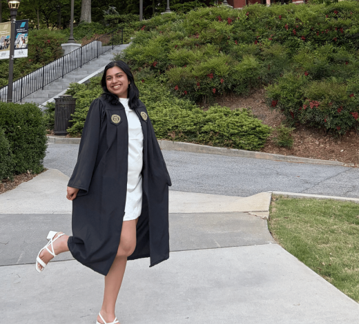 A graduate in cap and gown stands on the Georgia Tech campus with Tech Tower rising in the background, surrounded by trees and landscaped walkways.