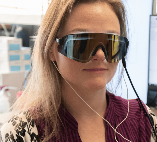 Person in a lab setting wearing large, wraparound goggles connected by a cable and earbud wires, seated in front of computer monitors and equipment, participating in a light and sound–based research experiment.