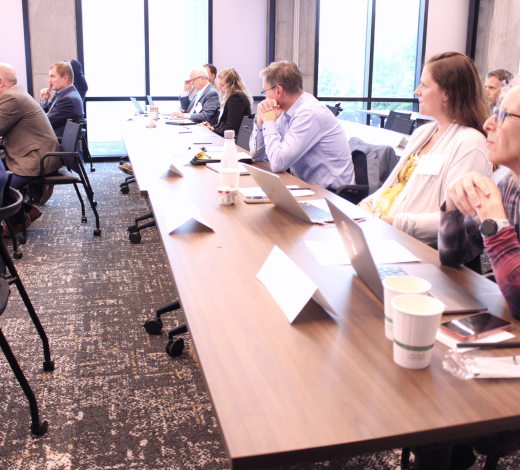 Audience members sit around a conference table in a bright meeting room, listening to a speaker holding a microphone; laptops, notebooks, and coffee cups are visible on the table near large windows.