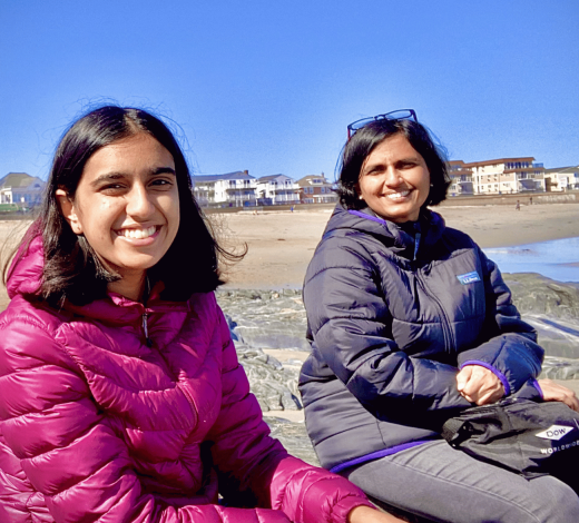 Two people in puffer jackets sit on rocks at a beach with beachfront homes and a clear blue sky in the background.