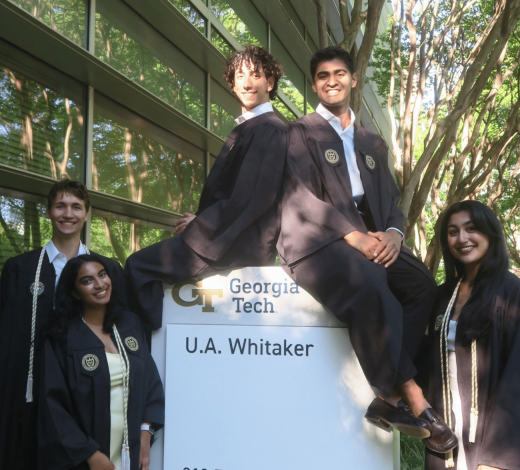 Five graduates wearing academic regalia pose around a Georgia Tech U.A. Whitaker building sign outdoors. Two graduates sit on top of the sign while three stand beside it. A glass building and leafy trees surround the group, with sunlight filtering through the branches. Visible text on the sign reads “Georgia Tech” and “U.A. Whitaker.”