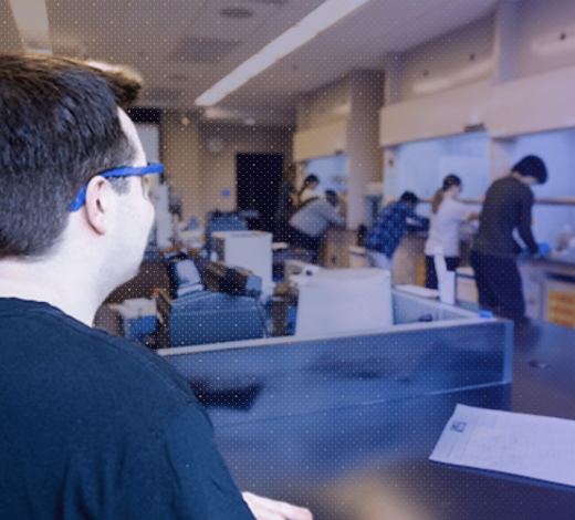 A student wearing safety glasses works in the foreground of a robotics lab as several other students work at equipment in the background.