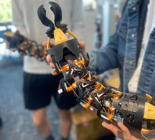 Close-up of a modular, snake-like robotic device with exposed wiring, sensors, and claw-like grippers at both ends being held by two people in a laboratory setting. The flexible robotic structure features black and orange components connected by segmented joints.