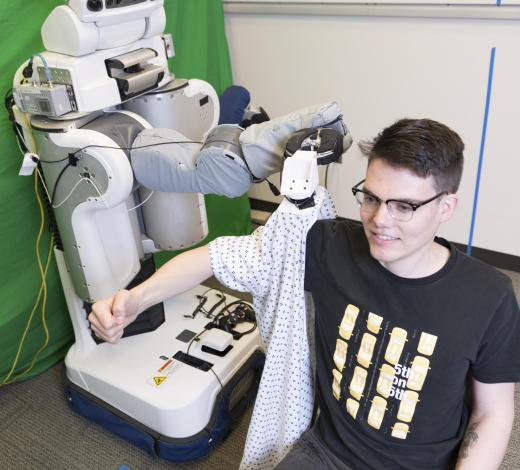 A PR2 robot puts a gown on Henry Clever, a member of the research team.