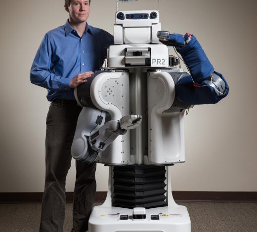 The blue cover on the arm of this robot protects an array of sensors that help it reach through the clutter of a typical home to perform personal care tasks. Charlie Kemp poses with the robot, which also holds an electric shaver. (Credit: Rob Felt)