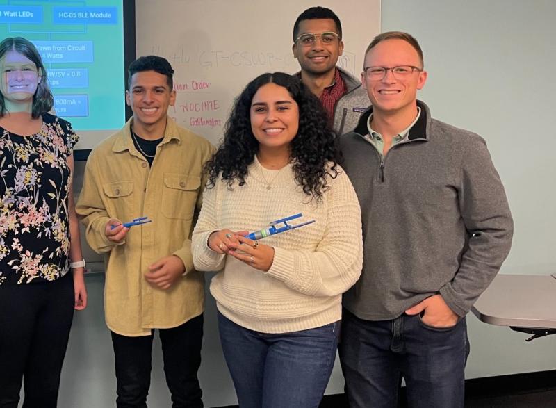 A photo of a group of individuals. From left to right: a male student, a female student, and a male professor. Another male student stands behind the female student.