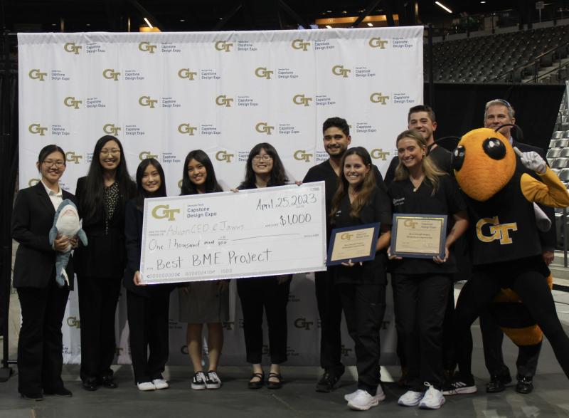 Photo of a group of nine students representing teams AdvanCED and Jawws holding a check for $1000. Two female students on the right side of the photo are holding small plaques. Next to the students on the far right of the photo is the Buzz mascot and Dr. James Stubbs, faculty advisor for BME Capstone.