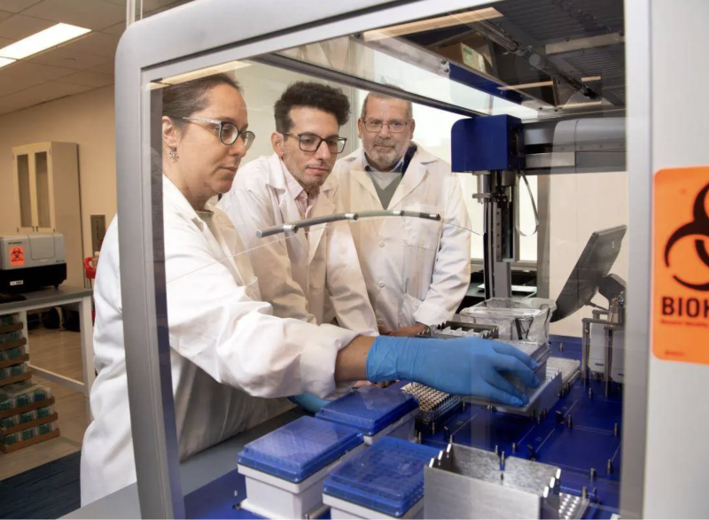 From left, Lorena Chaves, Jose Assumpcao, and Philip Santangelo are standing in a lab examining mRNA samples.