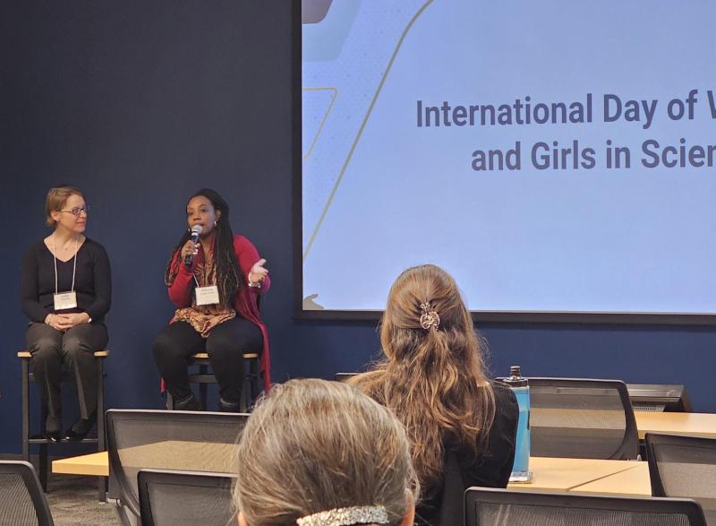 Photo of three women sitting at the front of a meeting room with a large screen that says "International Day of Women and Girls in Science." One woman who is African American, holds a mic while she is speaking.
