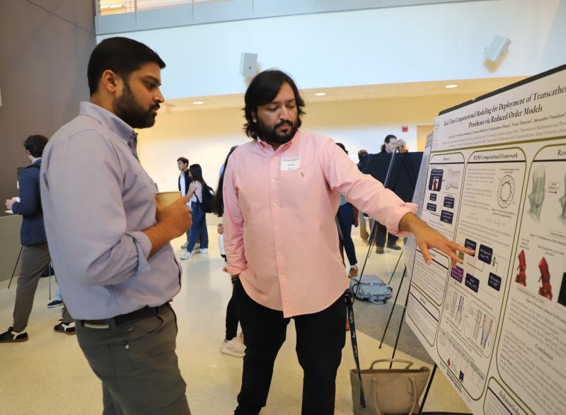 Photo of male student in a pink shirt in the center of the photo pointing to a poster of his research as another male student in a blue shirt on the left side of the photo looks on.