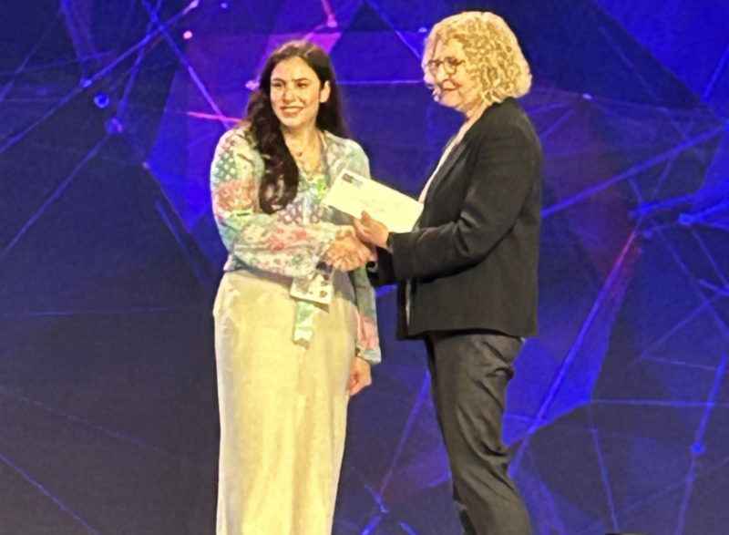 Close up photo of Retta El Sayed dressed in white standing on a stage being given an award by a female presenter.