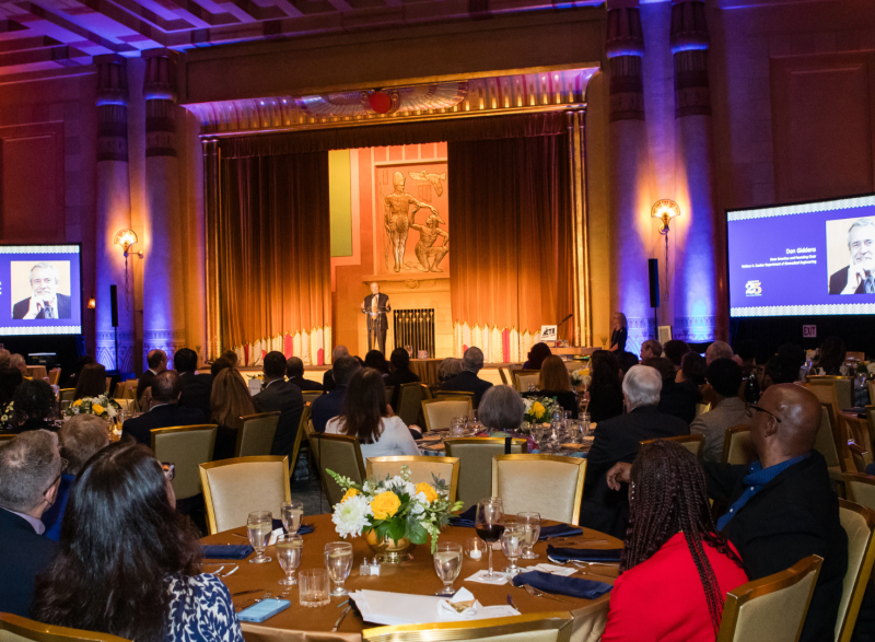 Photo of the Egyptian Ballroom at the Fox Theatre Atlanta looking toward the stage. In the foreground are people sitting in banquet chairs at tables looking at the speaker. In the background is the stage and speaker.