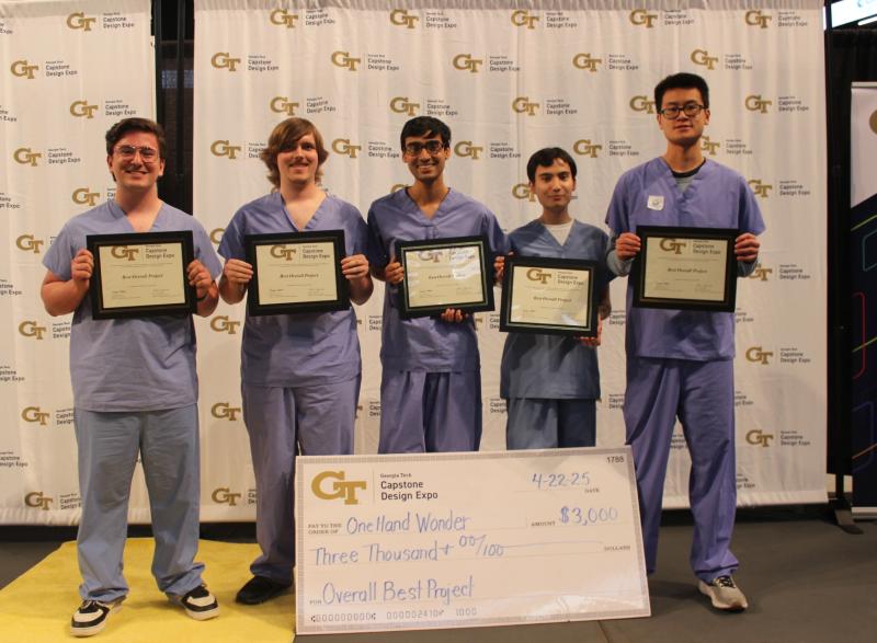 Five male students from Capstone team One-Hand Wonder holding gold plaques for winning first place at Capstone Expo. A large check fro $3,000 sits on the floor in front of them.