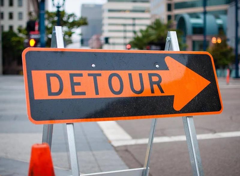 Photo of a detour road sign that has an orange border and an orange arrow with the words "DETOUR" in black inside of the arrow