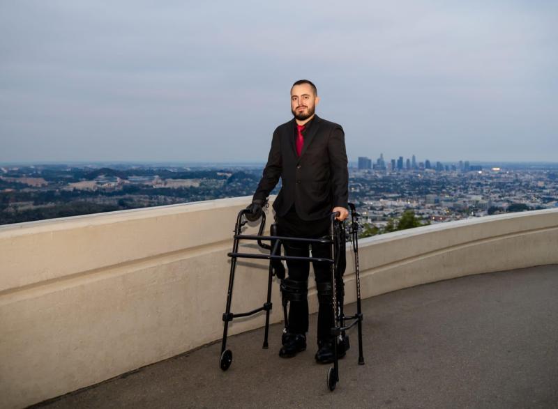 Photo of Coulter BME Ph.D. student Ignacio Montoya, standing on some sort of building rooftop with the skyline of Los Angeles behind him in the background. He is standing with the use of a walker.
