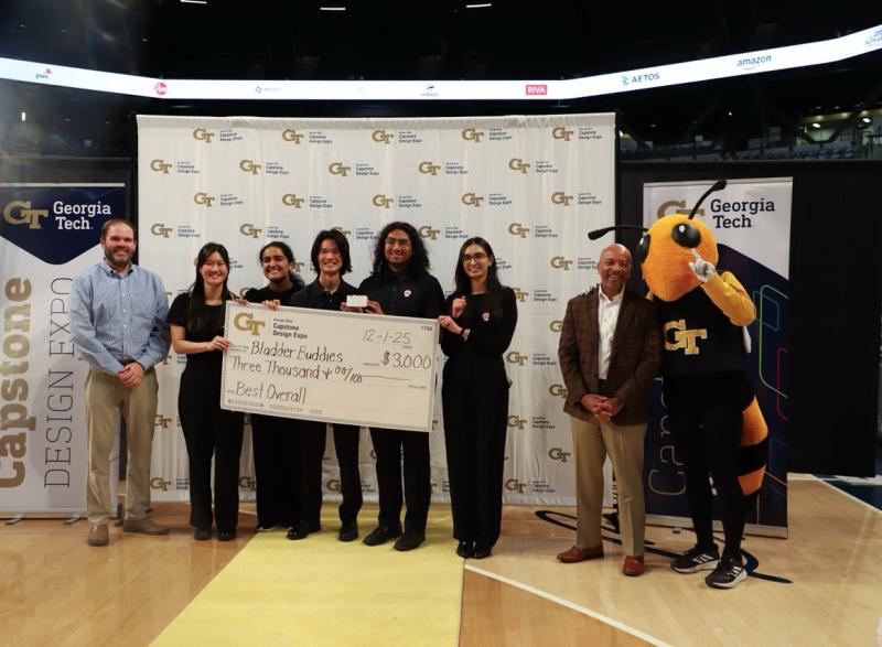 A photo of a team of students in black scrubs standing in front of a step and repeat banner holding a big check award while their faculty advisor stands to the left of them and the Georgia Tech Buzz mascot stands to the right of them next to the provost.