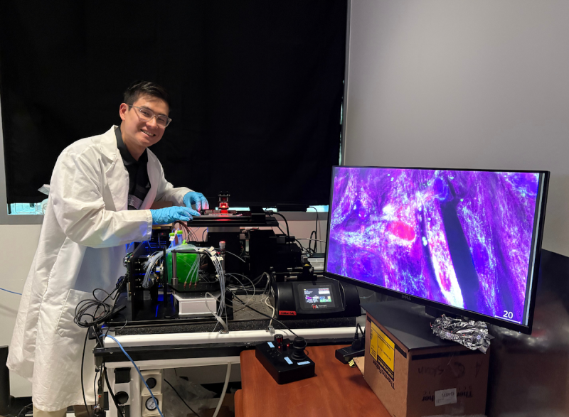 A person wearing a white lab coat and blue gloves is working at a laboratory station equipped with advanced imaging equipment. The setup includes multiple tubes, wires, and containers connected to a black optical device. A large monitor on the right displays a vivid, high-resolution microscopic image in shades of purple and pink, showing tissue structures. 