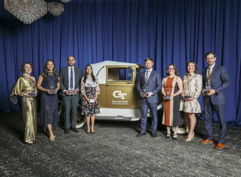A group of people stand on a stage in front of a vintage Georgia Tech Ramblin’ Wreck car, each holding a glass award, with blue draping and chandeliers in the background.