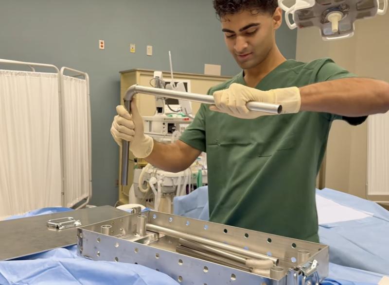 A person wearing surgical scrubs and gloves uses a metal tool to work with a stainless‑steel medical device or instrument tray in a clinical environment. Medical equipment and curtains are visible in the background.