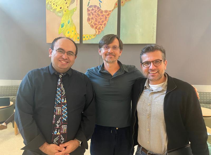 Three people stand side by side indoors for a group photo, with colorful scientific artwork displayed on the wall behind them