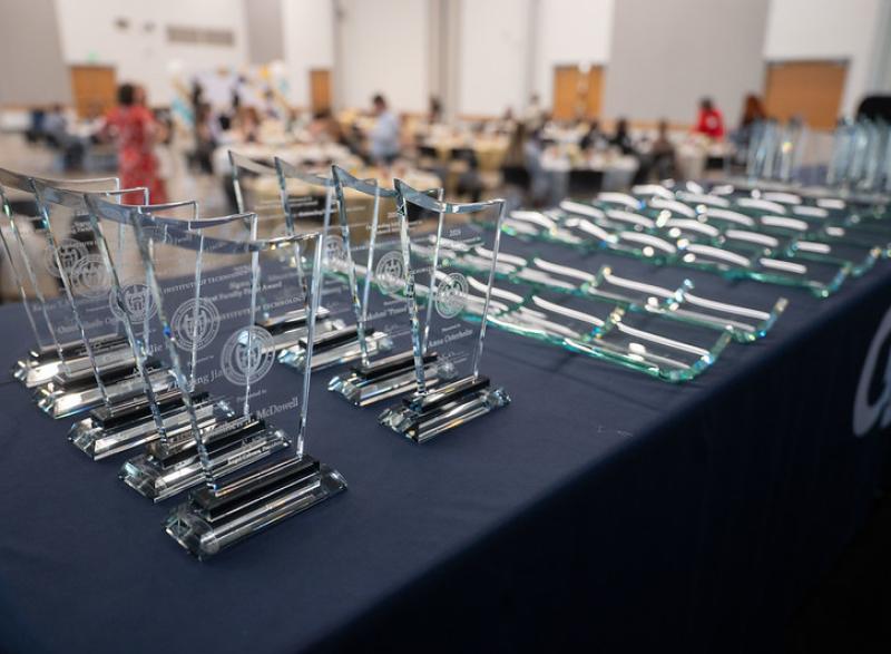 A row of clear glass award plaques arranged on a table in the foreground, with more plaques laid flat beside them and a blurred event setup in the background.