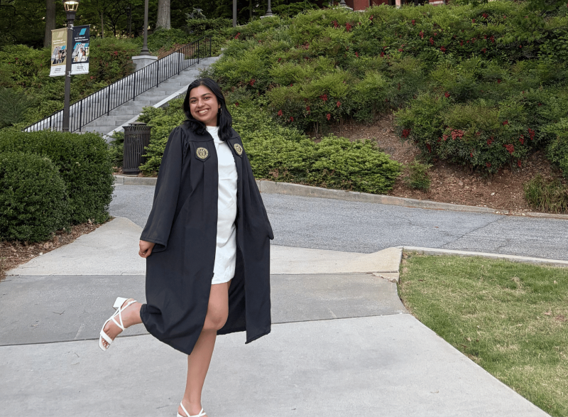 A graduate in cap and gown stands on the Georgia Tech campus with Tech Tower rising in the background, surrounded by trees and landscaped walkways.