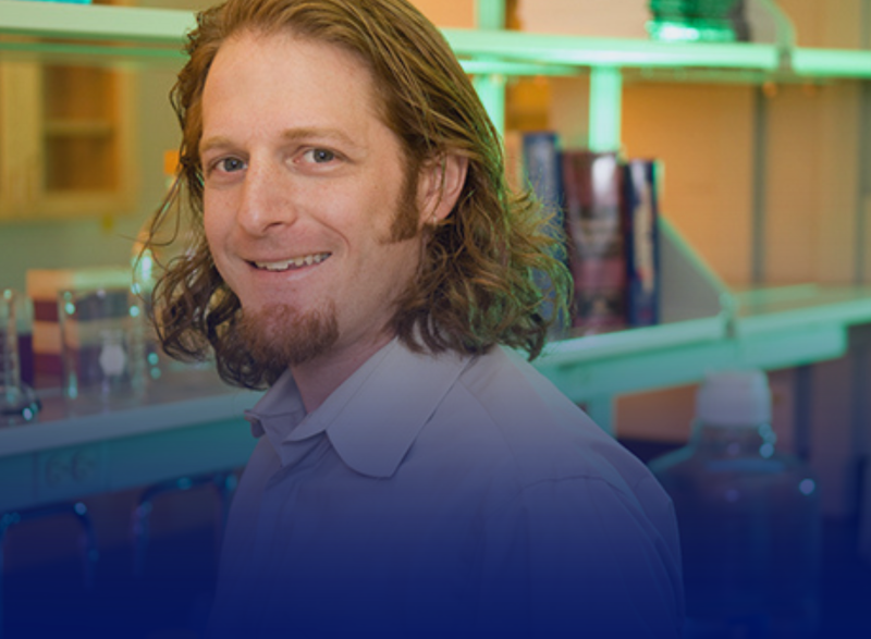 Smiling person with shoulder-length hair posing in a laboratory setting, with shelves of glass containers and equipment visible in the background.