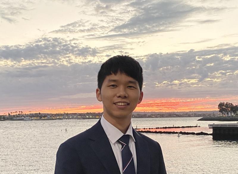 Person in a suit standing by the water at sunset, with a cloudy sky and shoreline in the background.