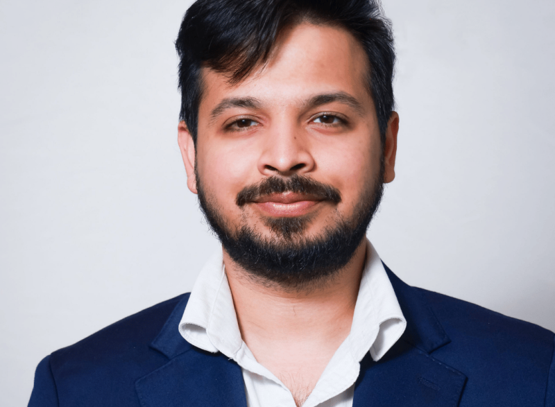 Close-up studio headshot of a person with short dark hair and a trimmed beard, wearing a navy blazer and white collared shirt against a light gray background.