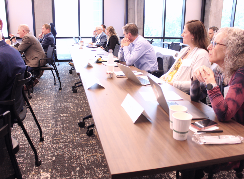 Audience members sit around a conference table in a bright meeting room, listening to a speaker holding a microphone; laptops, notebooks, and coffee cups are visible on the table near large windows.