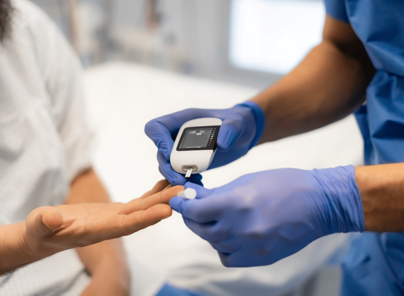 Healthcare worker wearing blue gloves uses a handheld glucose meter to check a patient’s blood sugar from a finger prick.