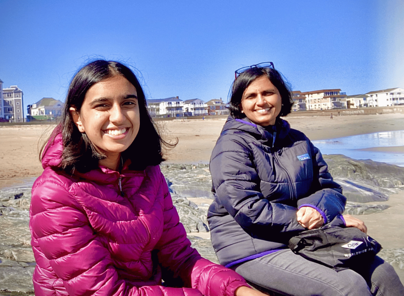Two people in puffer jackets sit on rocks at a beach with beachfront homes and a clear blue sky in the background.