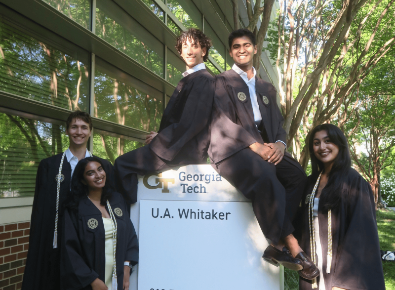 Five graduates wearing academic regalia pose around a Georgia Tech U.A. Whitaker building sign outdoors. Two graduates sit on top of the sign while three stand beside it. A glass building and leafy trees surround the group, with sunlight filtering through the branches. Visible text on the sign reads “Georgia Tech” and “U.A. Whitaker.”