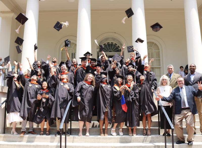 Graduates of the MBID program in Coulter BME are standing on the steps of the Historic Academy of Medicine with their caps and gowns on tossing their caps in the air. The department chair, two faculty, and the program director or standing on the right side of the steps. 