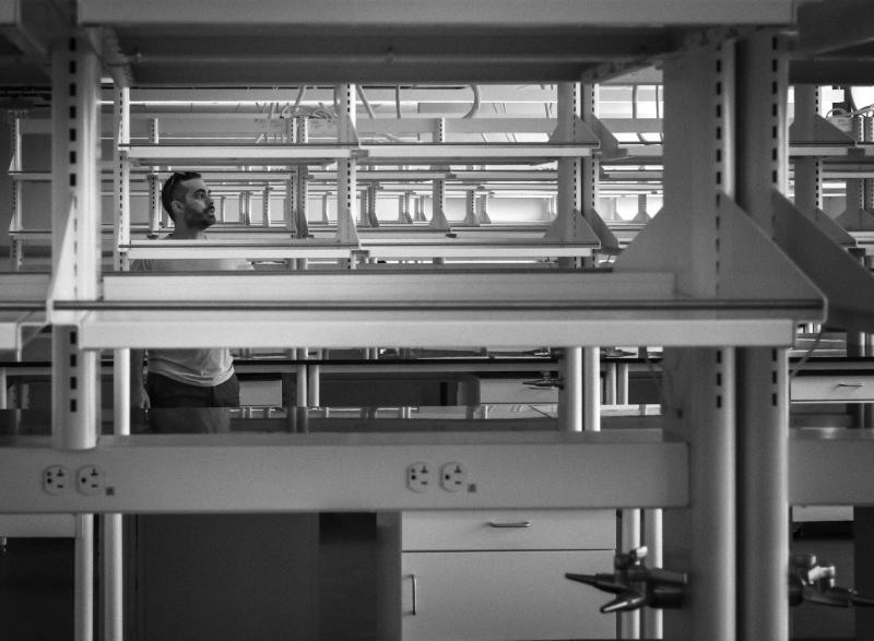 A person stands between rows of laboratory benches and shelving in a black-and-white research lab photograph.