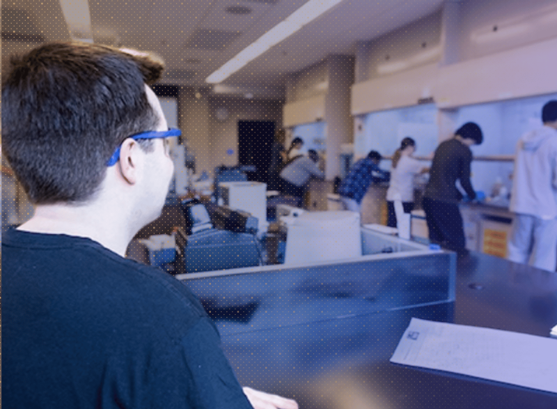 A student wearing safety glasses works in the foreground of a robotics lab as several other students work at equipment in the background.