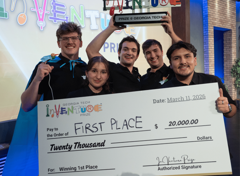 Five members of the winning Georgia Tech InVenture Prize team pose onstage holding a large ceremonial check for $20,000 and a trophy labeled “Prize @ Georgia Tech.” The oversized check reads “First Place” and “Winning 1st Place,” with the Georgia Tech InVenture Prize logo displayed prominently in the foreground. Colorful InVenture Prize signage appears in the background.