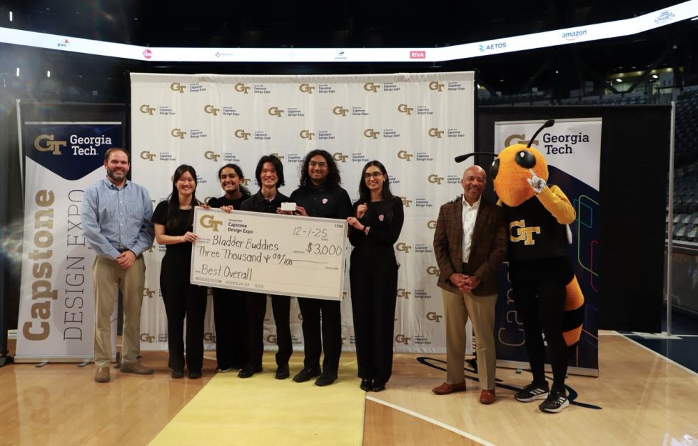 A photo of a team of students in black scrubs standing in front of a step and repeat banner holding a big check award while their faculty advisor stands to the left of them and the Georgia Tech Buzz mascot stands to the right of them next to the provost.