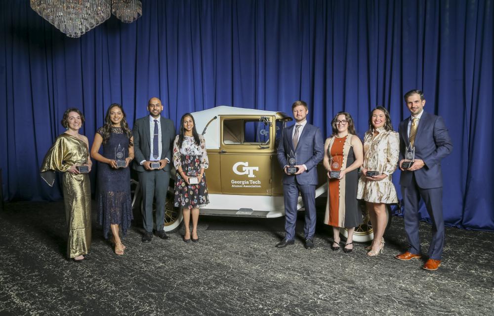 A group of people stand on a stage in front of a vintage Georgia Tech Ramblin’ Wreck car, each holding a glass award, with blue draping and chandeliers in the background.