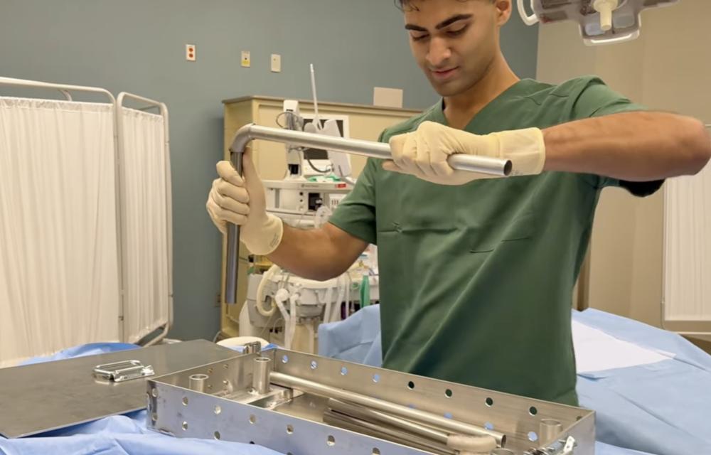 A person wearing surgical scrubs and gloves uses a metal tool to work with a stainless‑steel medical device or instrument tray in a clinical environment. Medical equipment and curtains are visible in the background.