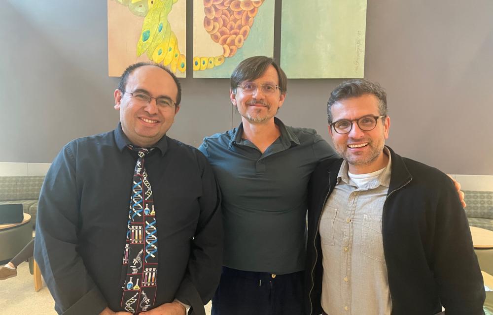 Three people stand side by side indoors for a group photo, with colorful scientific artwork displayed on the wall behind them