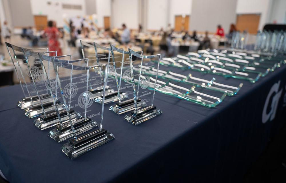 A row of clear glass award plaques arranged on a table in the foreground, with more plaques laid flat beside them and a blurred event setup in the background.