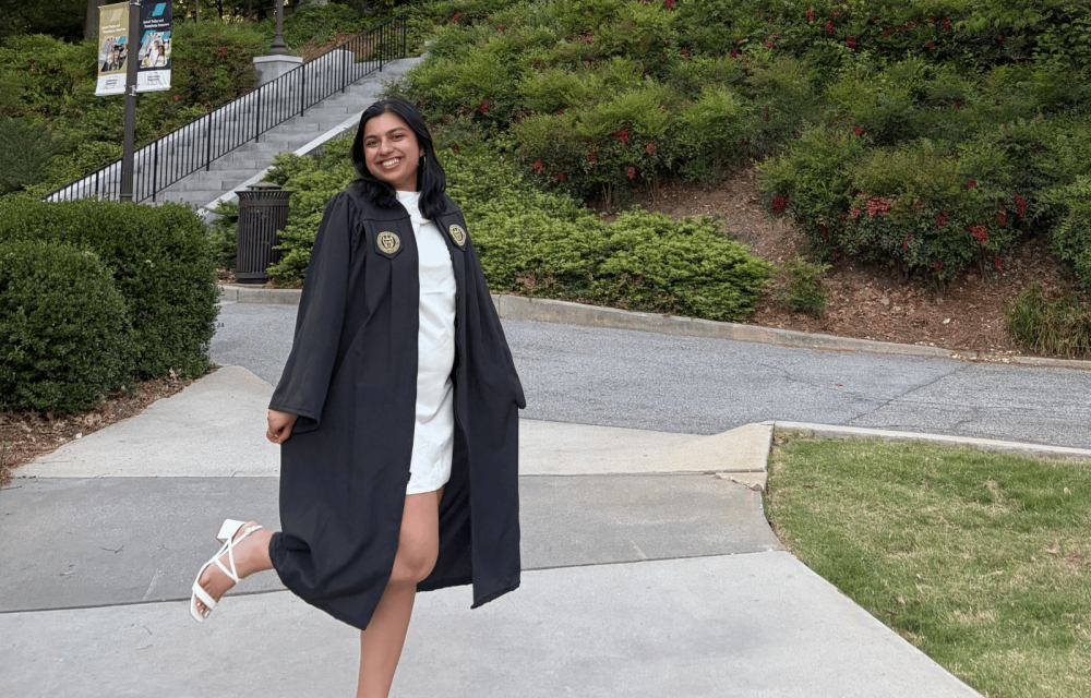 A graduate in cap and gown stands on the Georgia Tech campus with Tech Tower rising in the background, surrounded by trees and landscaped walkways.