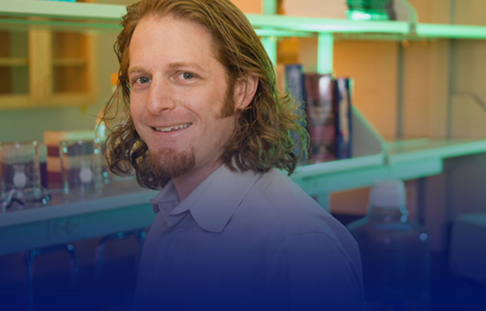 Smiling person with shoulder-length hair posing in a laboratory setting, with shelves of glass containers and equipment visible in the background.