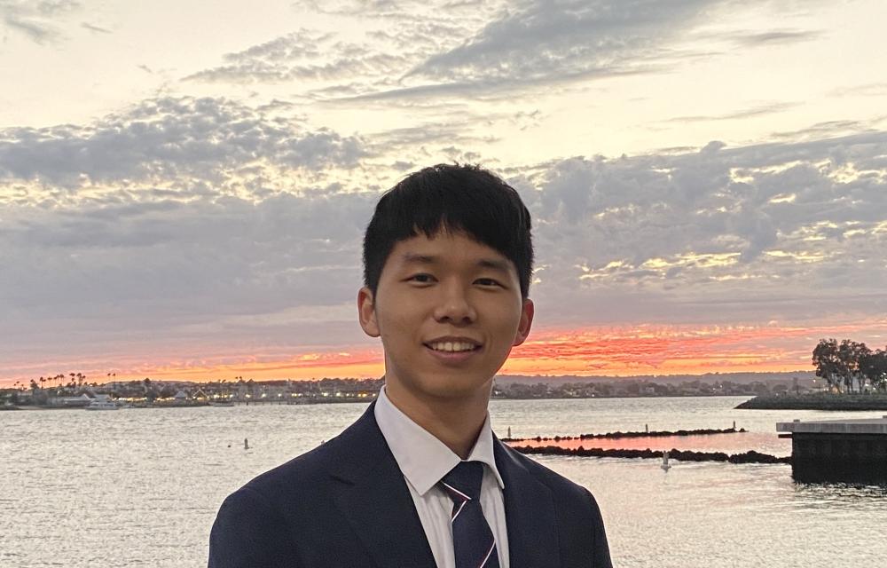 Person in a suit standing by the water at sunset, with a cloudy sky and shoreline in the background.