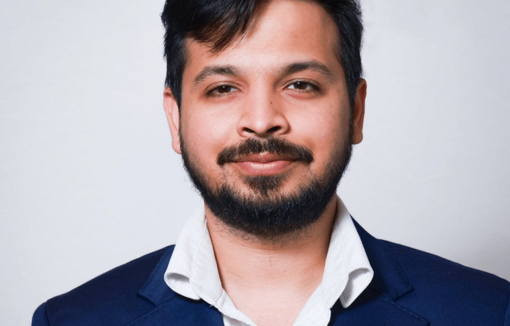 Close-up studio headshot of a person with short dark hair and a trimmed beard, wearing a navy blazer and white collared shirt against a light gray background.